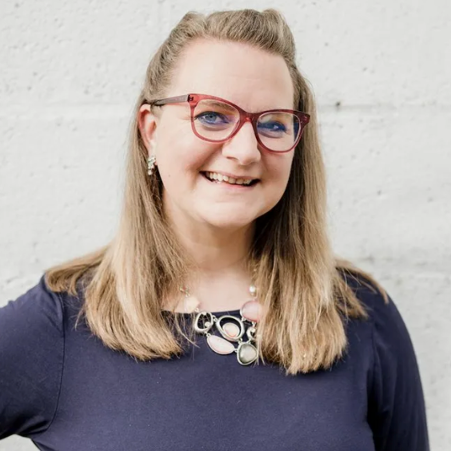 Beth Backes headshot wearing a blue dress and pink necklace in front of white brick wall